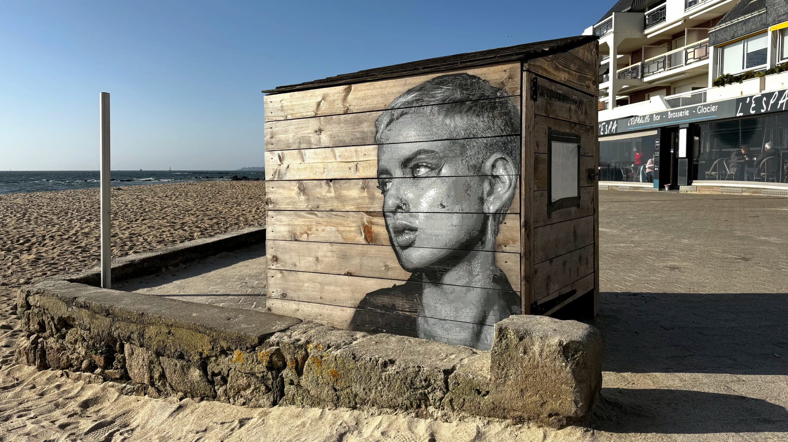 Portrait au pochoir d'une jeune femme aux cheveux courts. L'oeuvre a été réalisée par le street artiste Kelu Abstract en bord de mer et la jeune femme regarde vers le large.