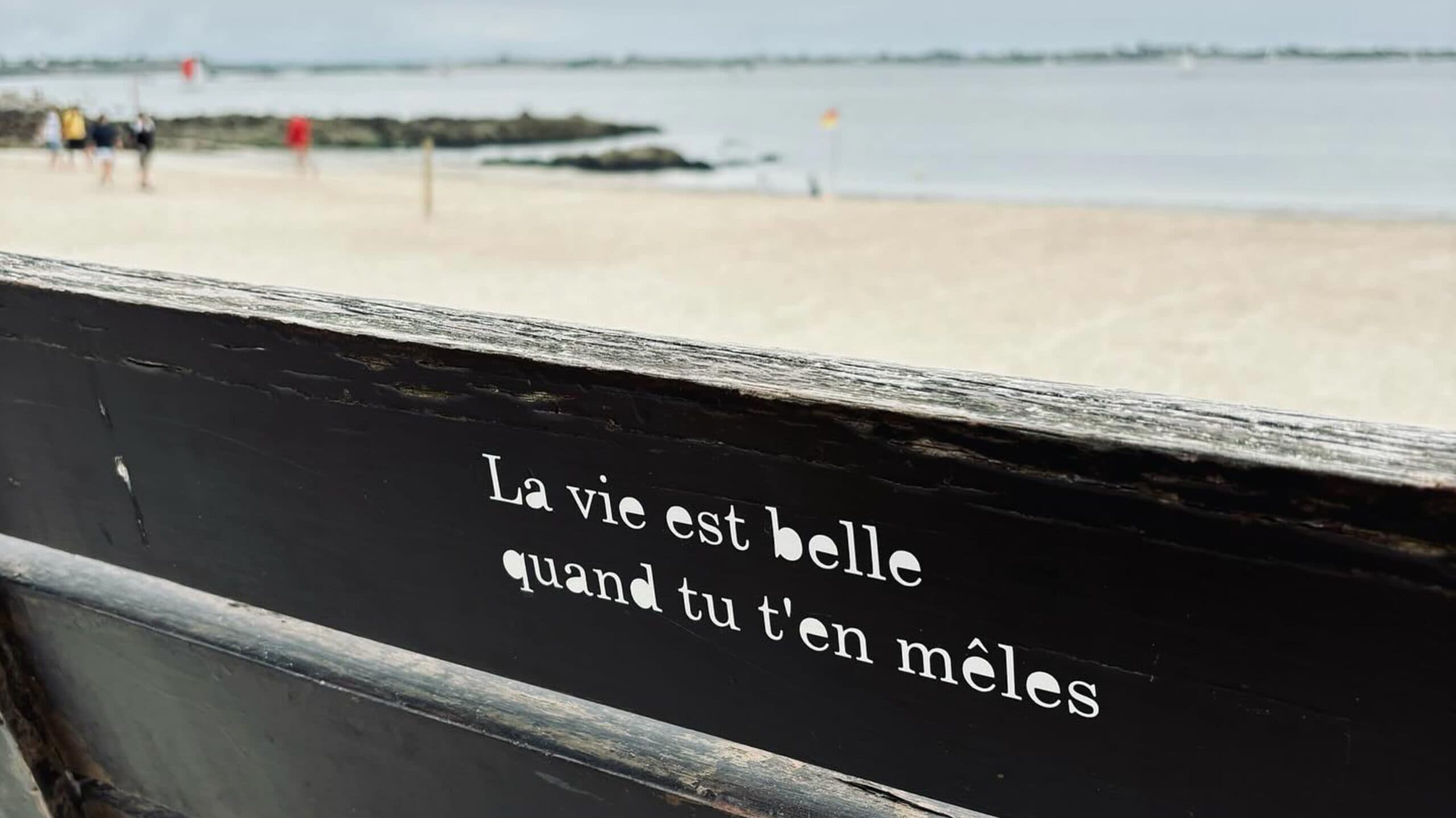 Aphorisme "La vie est belle quand tu t'en mêles" de Petite Poissone sur un banc de Larmor-Plage face à la mer.